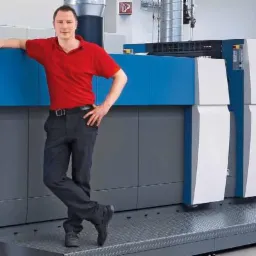 Mężczyzna oparty o niebiesko-białą maszynę drukarską Rapida 75 w hali drukarni. Widoczne panele sterowania i podajniki papieru.