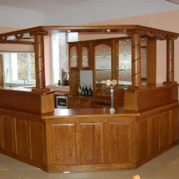 Drewniany bar z kolumnami i szklanymi półkami, oświetlony punktowo. W tle okno i szafki. Widoczny dystrybutor do piwa. Całość w ciepłej tonacji.