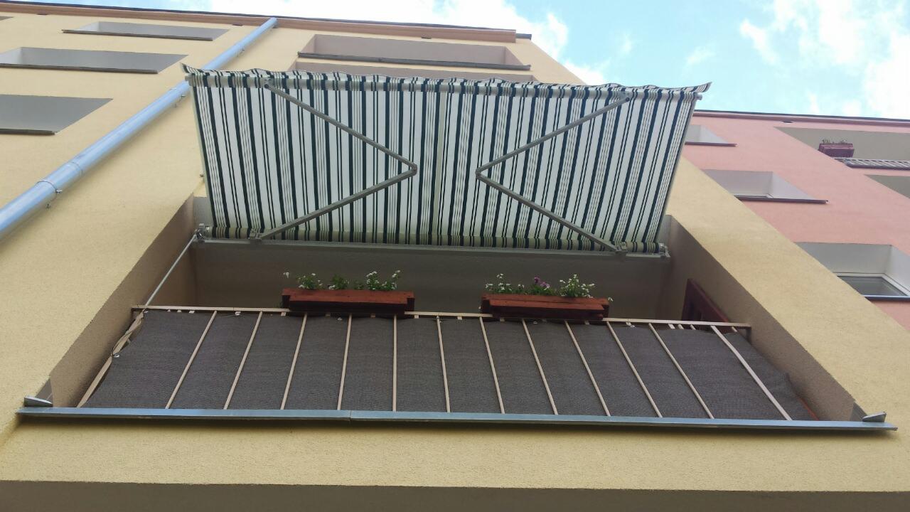 Zewnętrzna markiza balkonowa w biało-zielone paski, zamontowana nad balkonem z osłoną przeciwsłoneczną i doniczkami z kwiatami, widok z dołu na budynek mieszkalny.