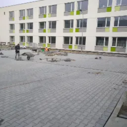 Prace brukarskie na placu przed budynkiem, widoczne ułożone fragmenty szarej kostki, pracownik z taczką i przygotowane materiały.