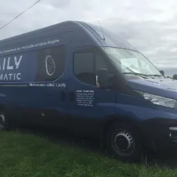 Niebieski samochód dostawczy Iveco Daily Hi-Matic z reklamą 8-stopniowej automatycznej skrzyni biegów, zaparkowany na trawie w pochmurny dzień.