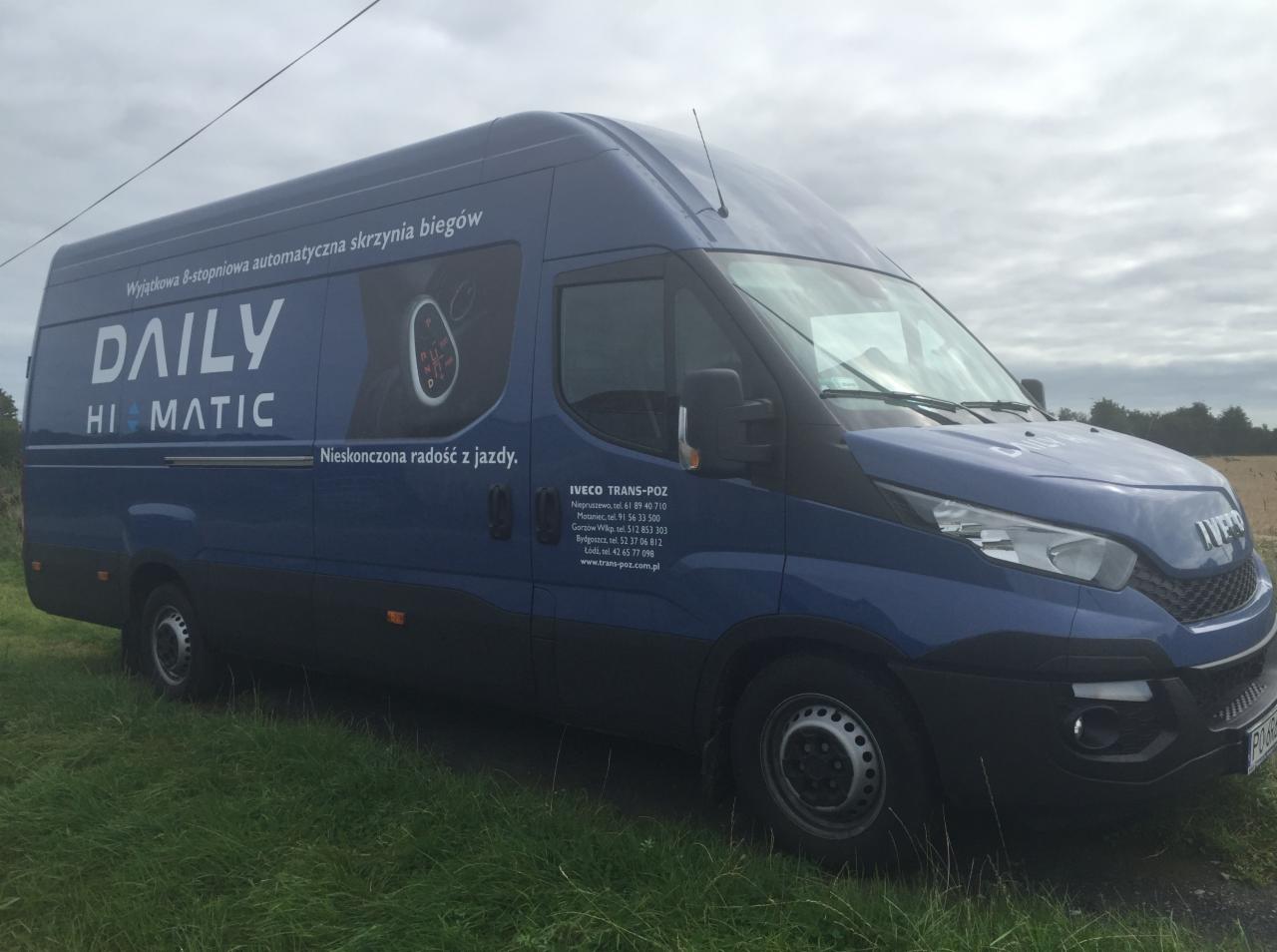 Niebieski samochód dostawczy Iveco Daily Hi-Matic z reklamą 8-stopniowej automatycznej skrzyni biegów, zaparkowany na trawie w pochmurny dzień.