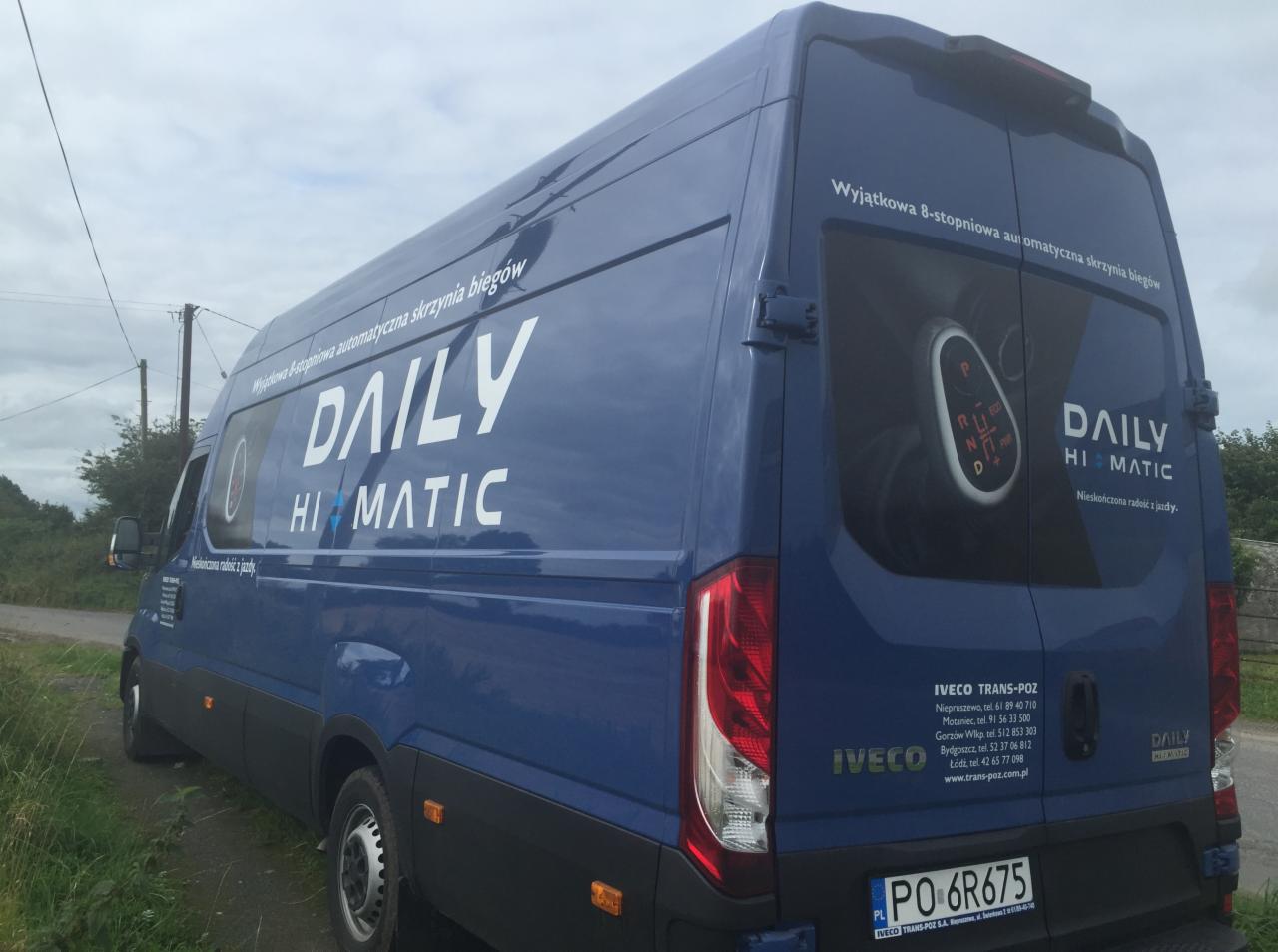 Niebieski samochód dostawczy Iveco Daily Hi-Matic z reklamą automatycznej skrzyni biegów, zaparkowany na poboczu drogi w pochmurny dzień.