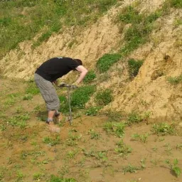 Pracownik w rękawicach wbija sondę glebową w zbocze porośnięte trawą i roślinnością, badanie gruntu na nierównym terenie.