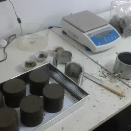 Próbki gruntu w metalowych cylindrach i pojemnikach, przygotowane do badań laboratoryjnych, w tle waga analityczna i akcesoria.