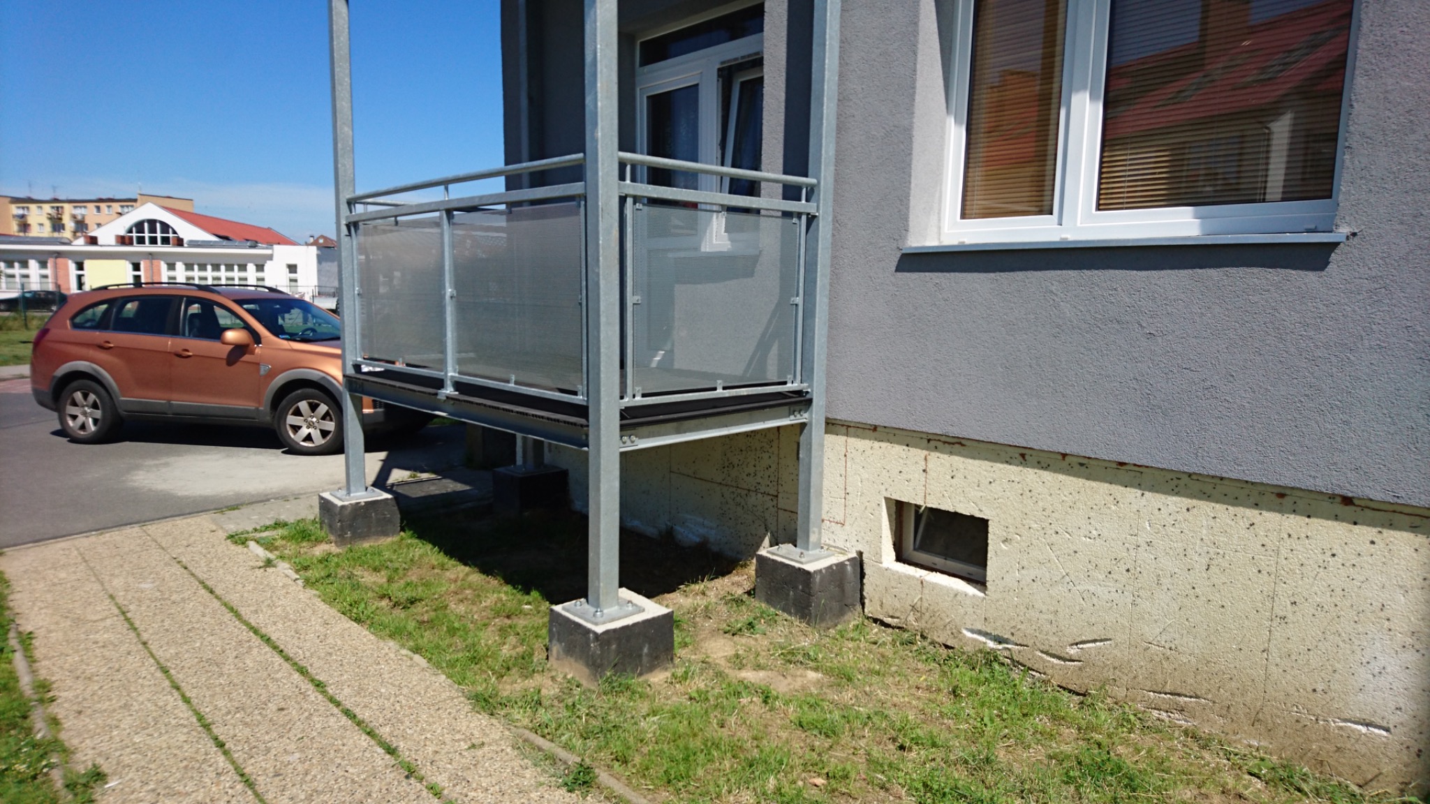 Zewnętrzny balkon ze stalową konstrukcją i ażurową balustradą, zamontowany przy szarym budynku mieszkalnym z widoczną elewacją i oknem z żaluzjami, obok chodnika i trawnika.