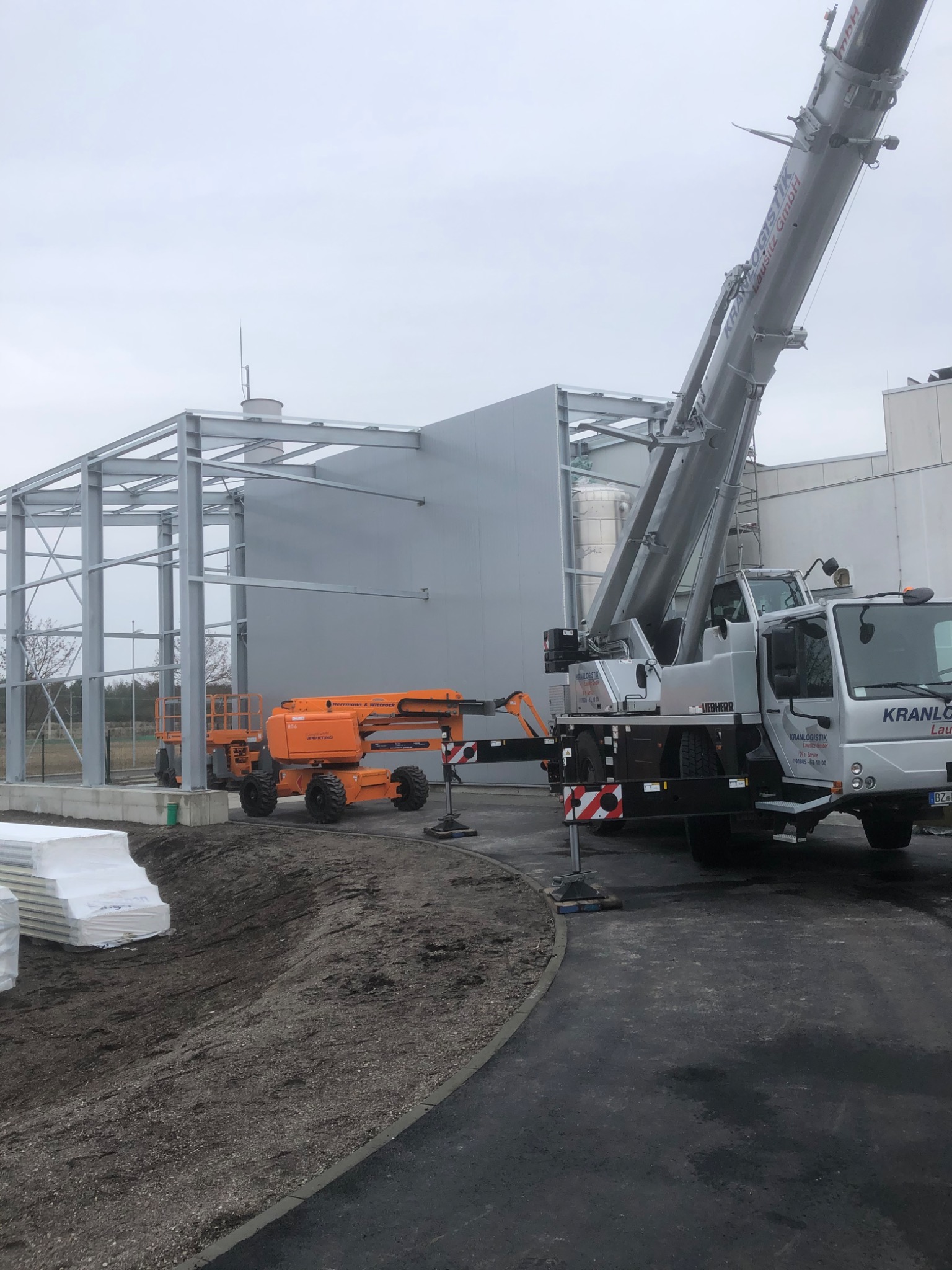 Wznoszenie stalowej konstrukcji hali z użyciem dźwigu Liebherr i podnośników koszowych Herrmann & Wittrock, widoczne płyty warstwowe czekające na montaż.