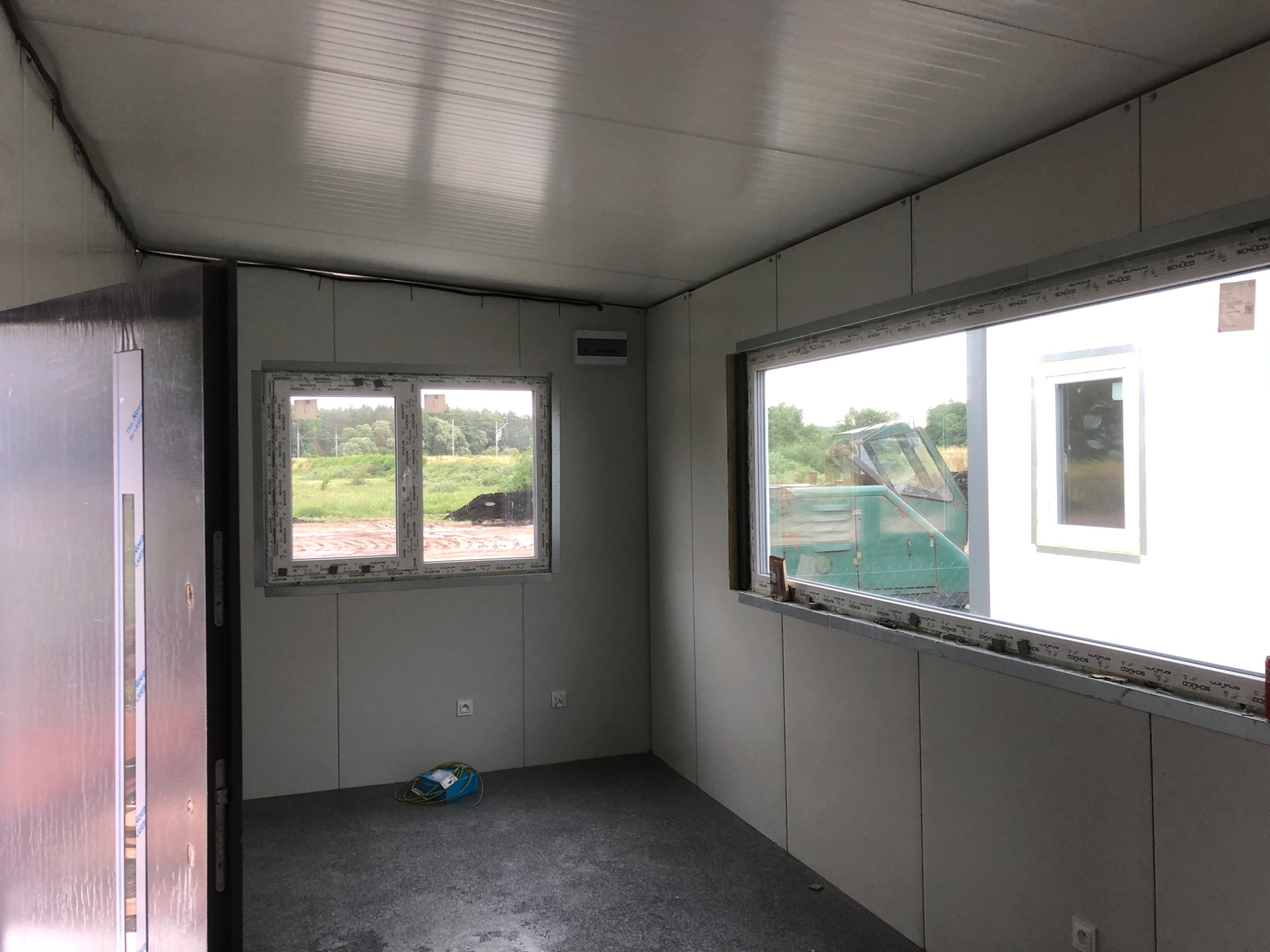 Wnętrze hali w trakcie budowy z widocznymi oknami, płytami warstwowymi na ścianach i suficie, oraz kablem elektrycznym na podłodze.