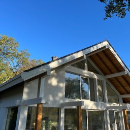 PBD Elewacje - Nowoczesny dom z jasną elewacją, dużymi oknami i widoczną drewnianą konstrukcją dachu, oświetlony słońcem, z zielonymi drzewami w tle.