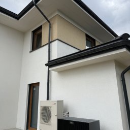 PBD Elewacje - Nowoczesny dom z białą, fakturowaną fasadą i beżowym akcentem, z czarnymi rynnami. Przy narożniku zainstalowano pompę ciepła Mitsubishi Electric i jednostkę Viessmann.