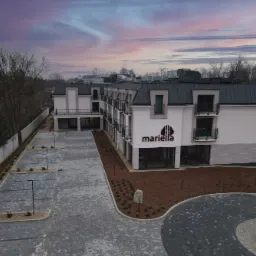 Widok z lotu ptaka na nowo wybudowany, trzypiętrowy budynek z elewacją w odcieniach bieli i szarości, z balkonami i parkingiem wyłożonym kostką brukową, pod błękitno-różowym niebem o zmierzchu.