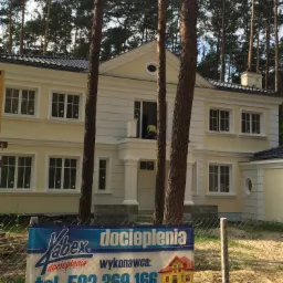 Żółty, dwupiętrowy dom z białymi ramami okiennymi i czarnym dachem, częściowo zasłonięty przez trzy drzewa, z reklamą firmy ociepleniowej na pierwszym planie, widoczny pracownik w oknie.