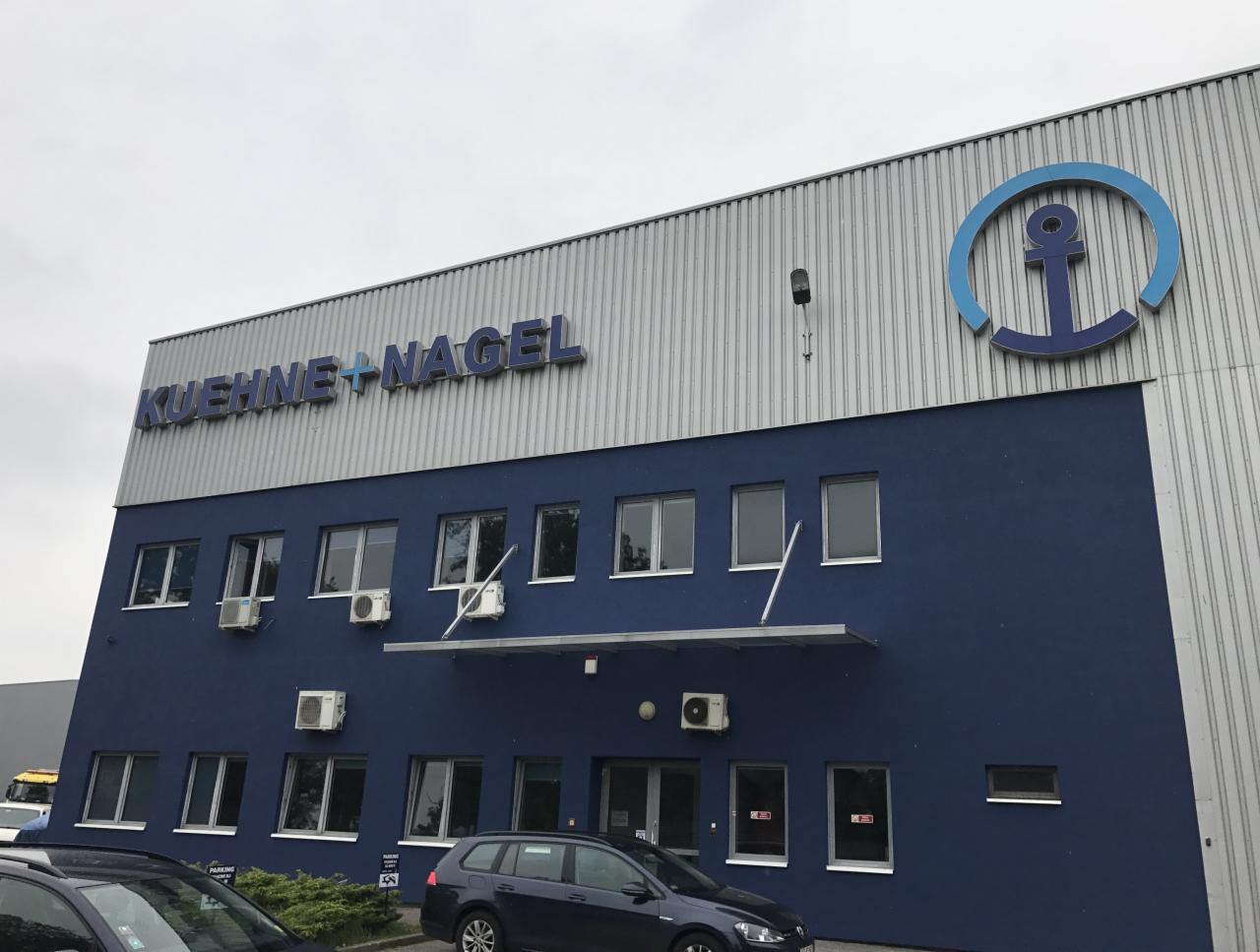 Dwupiętrowy budynek biurowy Kuehne+Nagel z niebieską elewacją i szarą, metalową górną częścią, zwieńczony logo firmy z kotwicą.