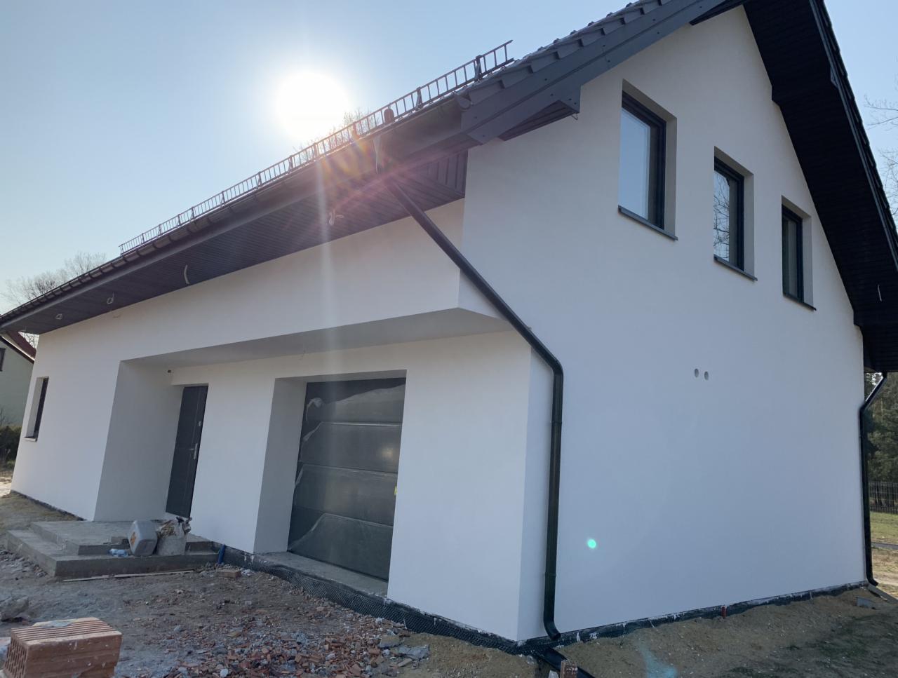 Świeżo otynkowana na biało elewacja domu jednorodzinnego z czarnymi oknami i dachem, widoczny teren budowy z cegłami i resztkami materiałów.