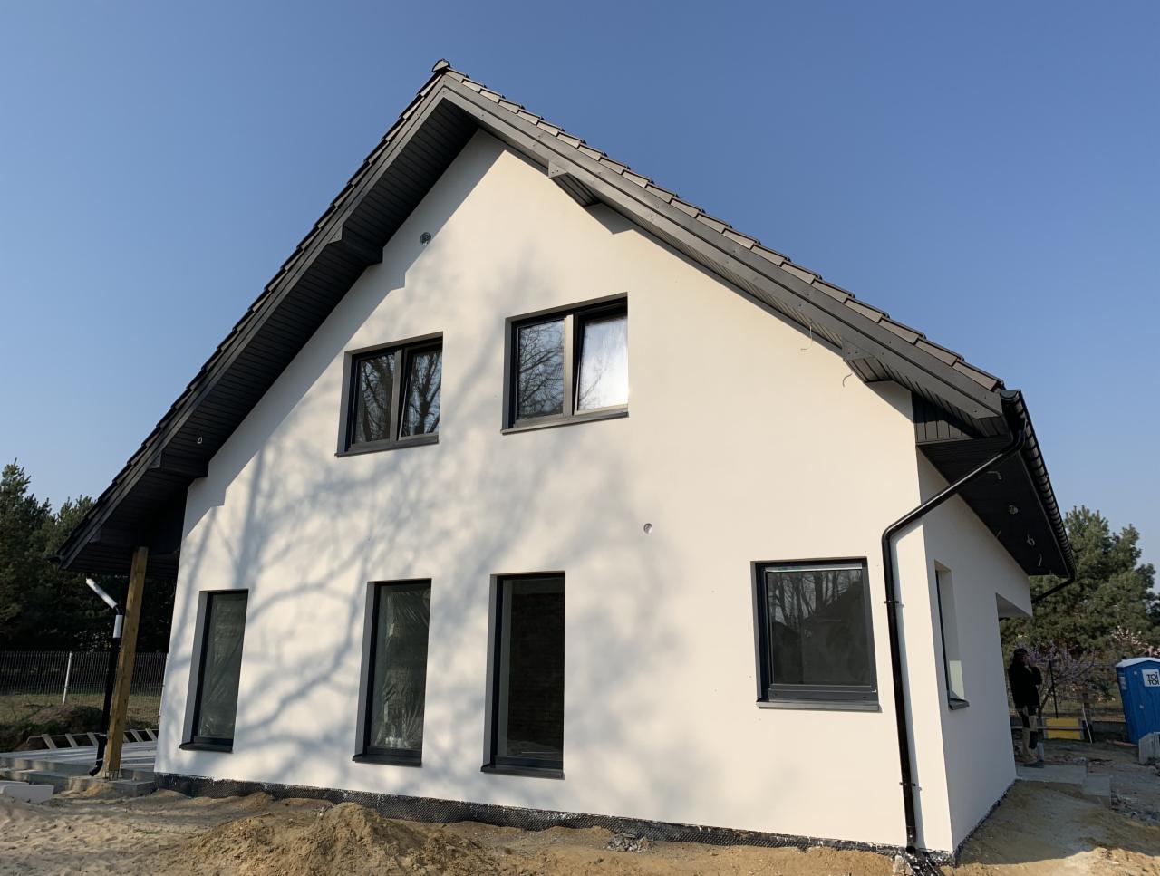 Nowoczesny dom jednorodzinny z jasną elewacją i ciemną stolarką okienną, widoczne ślady prac wykończeniowych wokół budynku, błękitne niebo w tle.