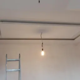 Wykończony sufit podwieszany z oświetleniem tymczasowym, widoczna drabina i drzwi w surowym wnętrzu.