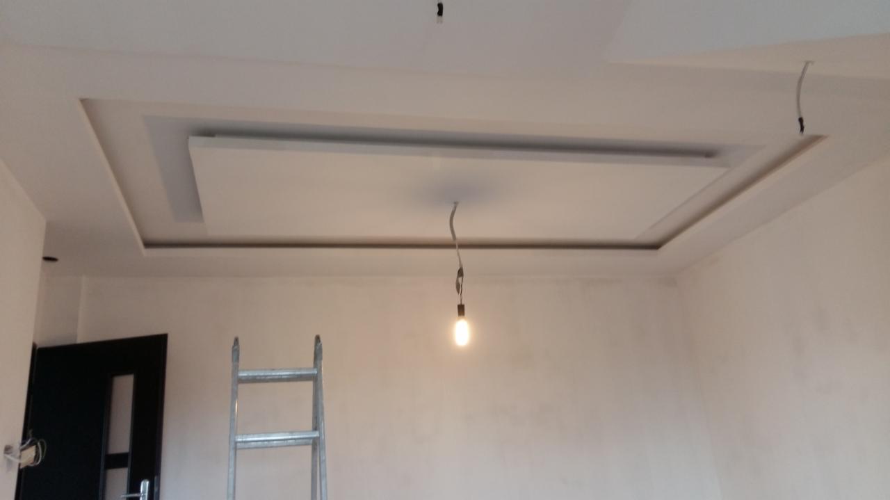 Wykończony sufit podwieszany z oświetleniem tymczasowym, widoczna drabina i drzwi w surowym wnętrzu.