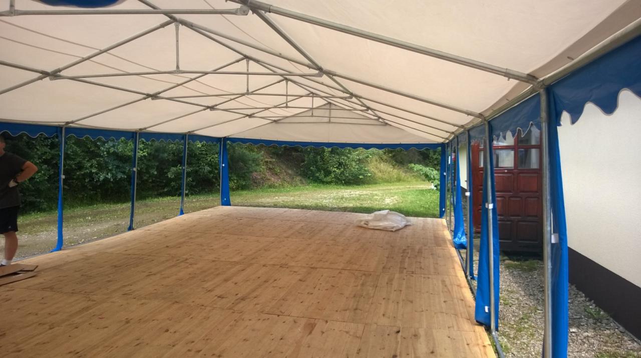 Wnętrze hali namiotowej 6x12m z drewnianą podłogą, niebieskimi obrzeżami i widoczną osobą w lewym rogu; tło z zielenią.