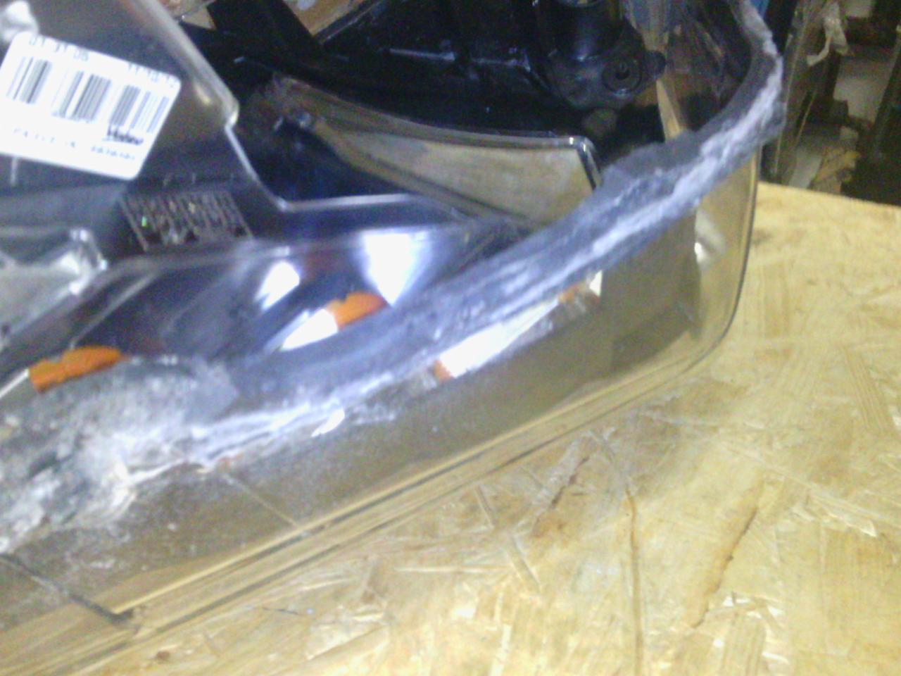 Uszkodzony reflektor samochodowy z widocznymi śladami naprawy, leżący na drewnianej powierzchni. Widoczne pęknięcia i ślady klejenia.