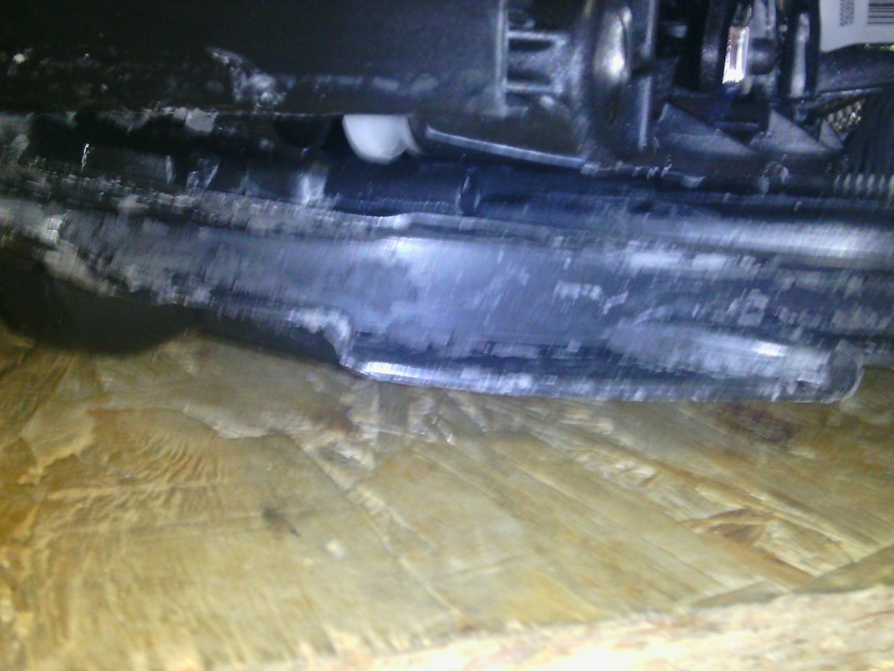 Nisko położony fragment podwozia pojazdu z widocznymi elementami konstrukcyjnymi i śladami eksploatacji, na tle drewnianej powierzchni.