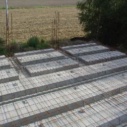Zbrojona płyta fundamentowa z pustaków szalunkowych na tle pola i zieleni. Widoczne pręty zbrojeniowe. Górny kadr.