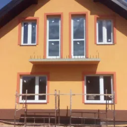 Żółta elewacja domu z białymi oknami, obramowana pomarańczową farbą. Na dole metalowe rusztowanie. Widoczne szpilki pod parapetem. Dom w trakcie wykańczania.