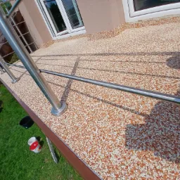 Balkon wykończony żywiczną posadzką z drobnymi kamyczkami, stalowa balustrada, widoczne puszki z farbą i wiadro na trawie.