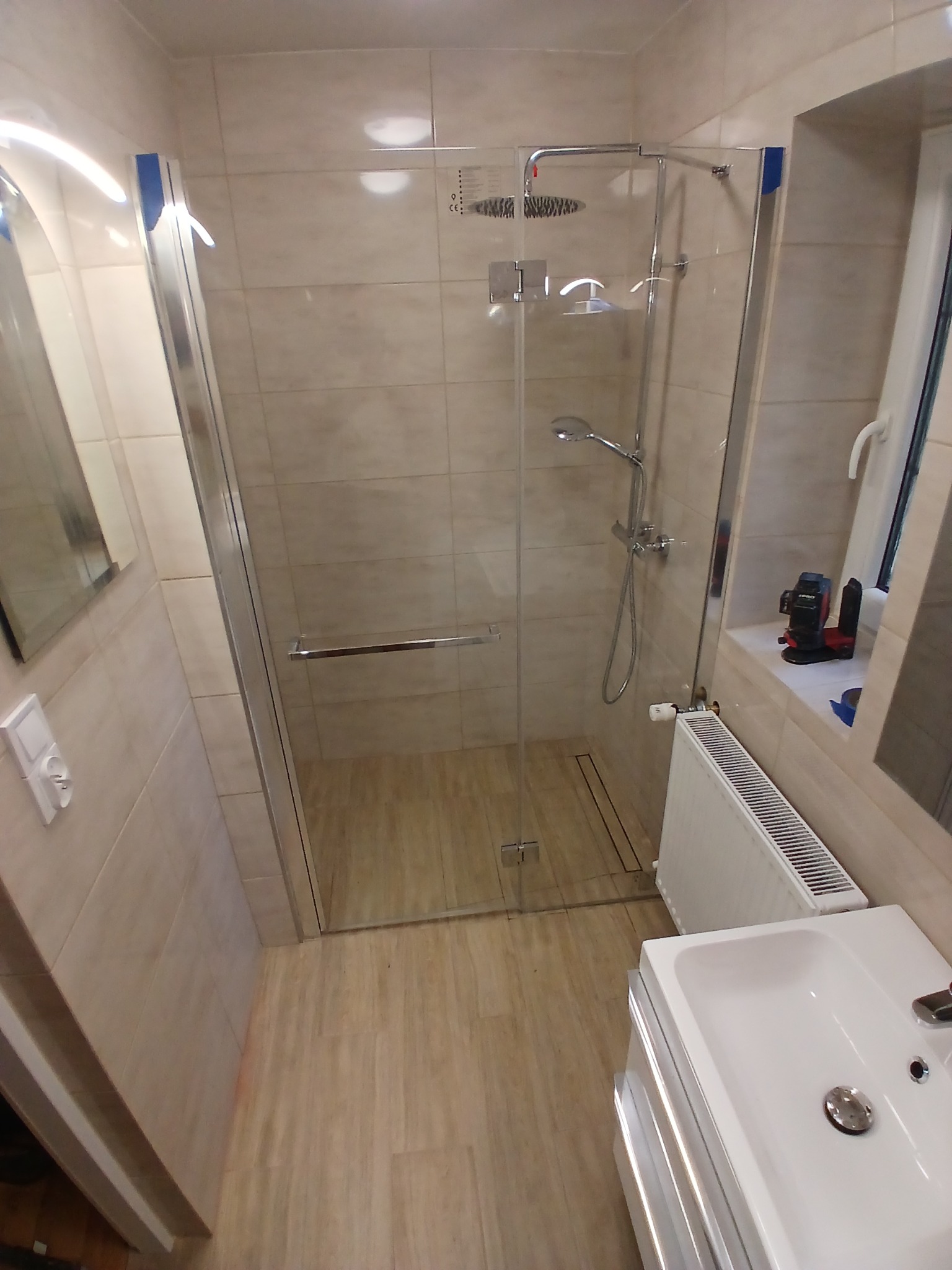 Wyremontowana łazienka z kabiną prysznicową typu walk-in, beżowe płytki na ścianach i podłodze imitujące drewno, biały grzejnik i umywalka, poziomica laserowa na parapecie okna.