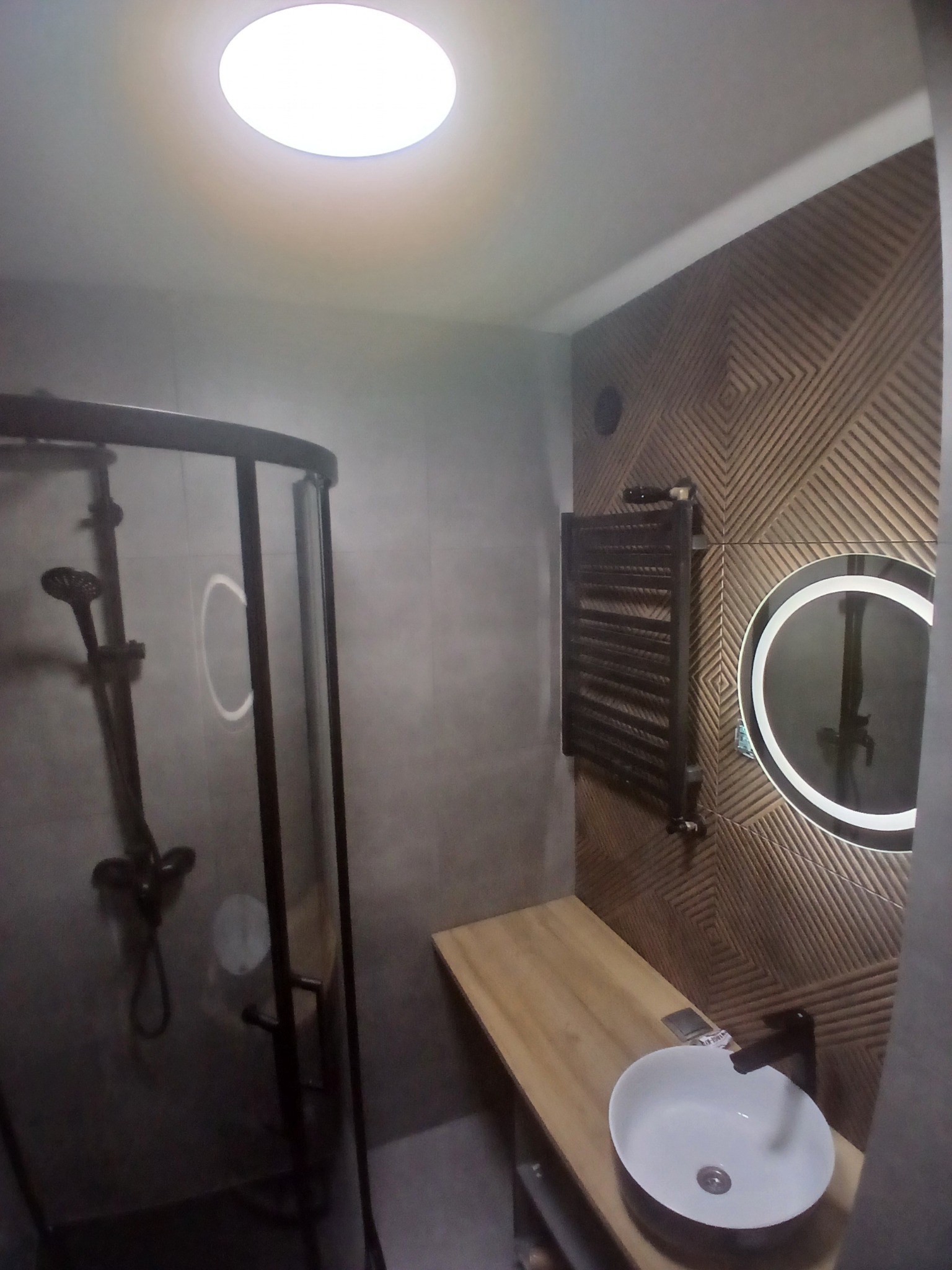 Nowoczesna łazienka z kabiną prysznicową z czarną armaturą, drewnianym blatem z białą umywalką, grzejnikiem drabinkowym i okrągłym lustrem z podświetleniem LED na dekoracyjnej ścianie z wzorem...