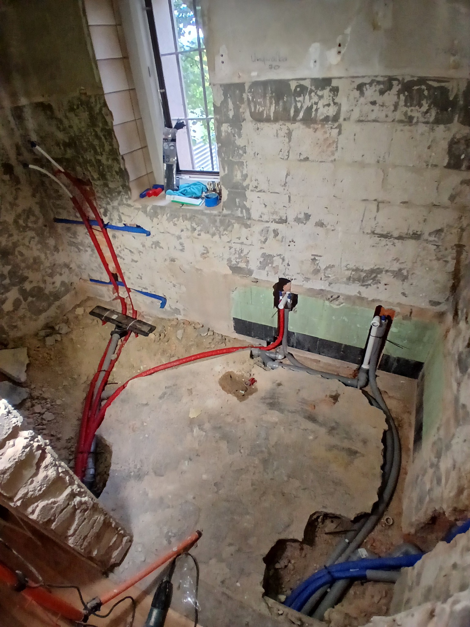 Surowa łazienka w trakcie remontu: widoczne instalacje wodne w czerwonych i szarych rurach, ściany z odsłoniętą cegłą i fragmentami starej glazury, okno z widokiem na zieleń.