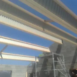 Konstrukcja dachu z drewnianymi belkami i blachą trapezową, widok od dołu na tle błękitnego nieba, częściowo widoczny mur z bloczków betonowych i rusztowanie.