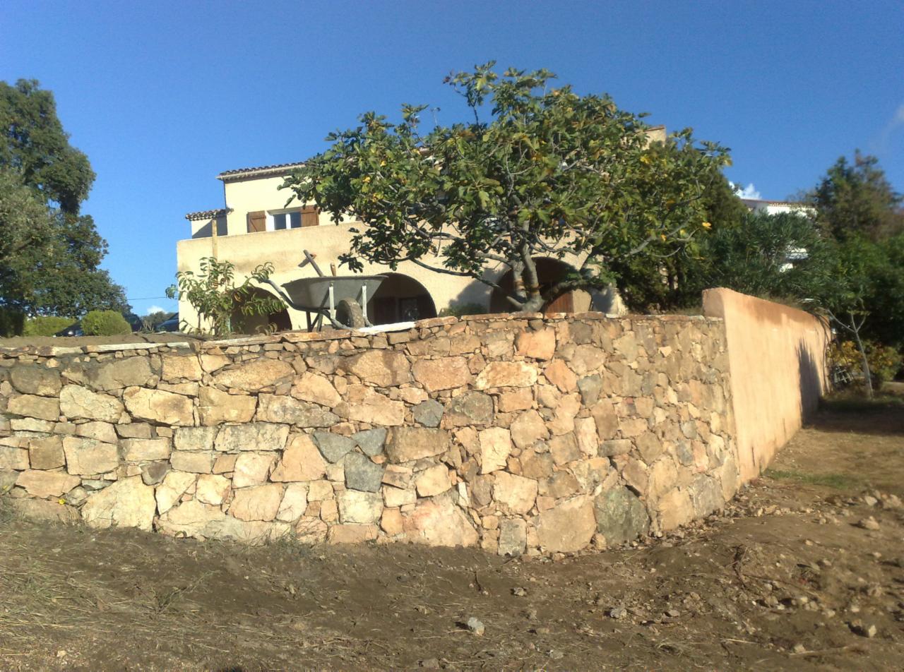 Dwupiętrowy dom z beżową elewacją, widoczny zza kamiennego muru, z drzewem owocowym na pierwszym planie i taczką zaparkowaną pod łukowym podcieniem.