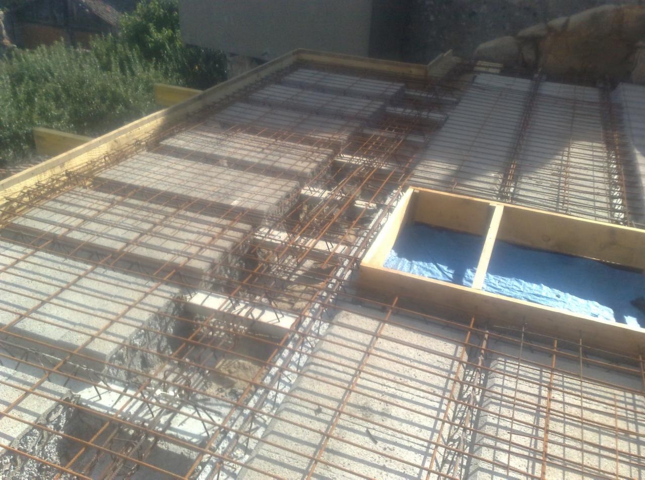 Zbrojenie stropu betonowego z drewnianym szalunkiem i widoczną niebieską folią hydroizolacyjną w otworze, przygotowane do zalania betonem, z widocznym fragmentem zieleni w tle.