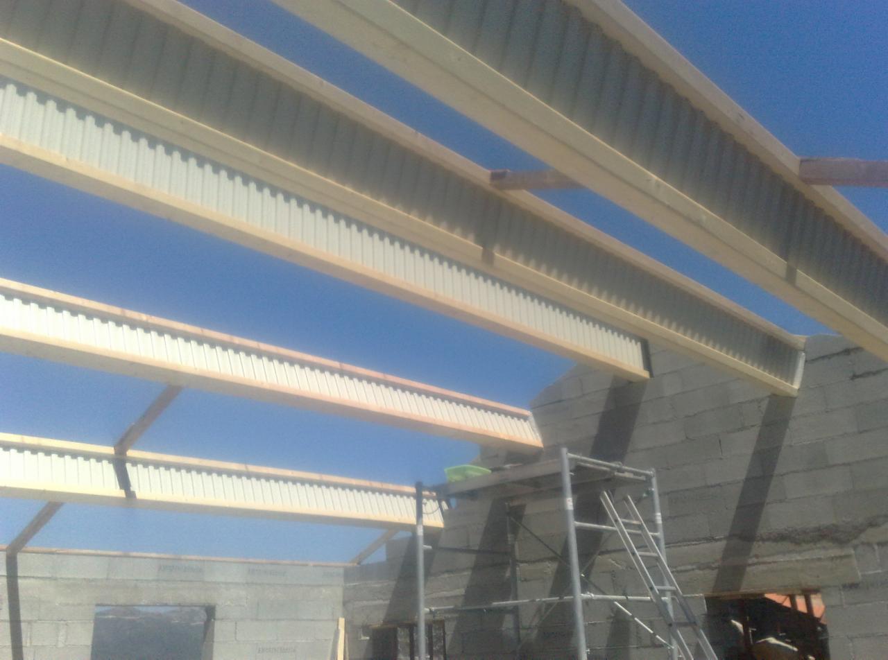 Konstrukcja dachu z drewnianymi belkami i blachą trapezową, widok od dołu na tle błękitnego nieba, częściowo widoczny mur z bloczków betonowych i rusztowanie.