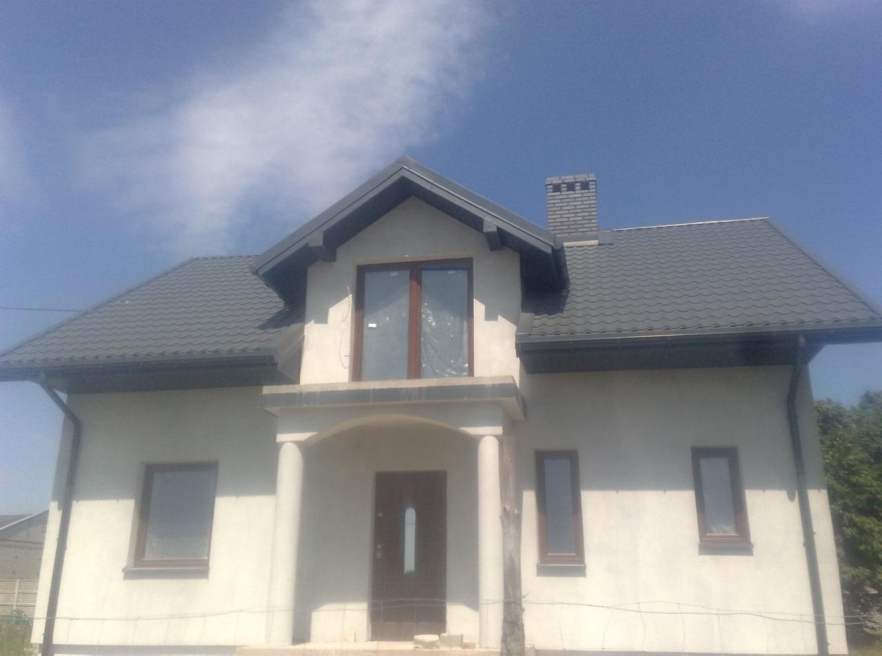 Niedokończony dom murowany z ciemnoszarą blachodachówką, widocznym kominem z cegły i oknami zabezpieczonymi folią.