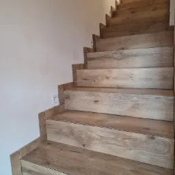 Schody wyłożone płytkami imitującymi drewno, widoczne plastikowe krzyżyki dystansowe, białe ściany w tle.