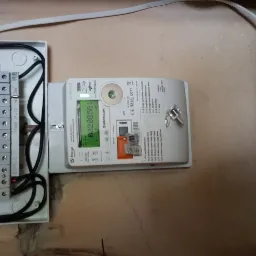 Otwarta skrzynka elektryczna z bezpiecznikami i licznikiem energii elektrycznej firmy Sagemcom, widoczne przewody i elementy instalacji na tle ściany z widocznymi śladami po malowaniu.
