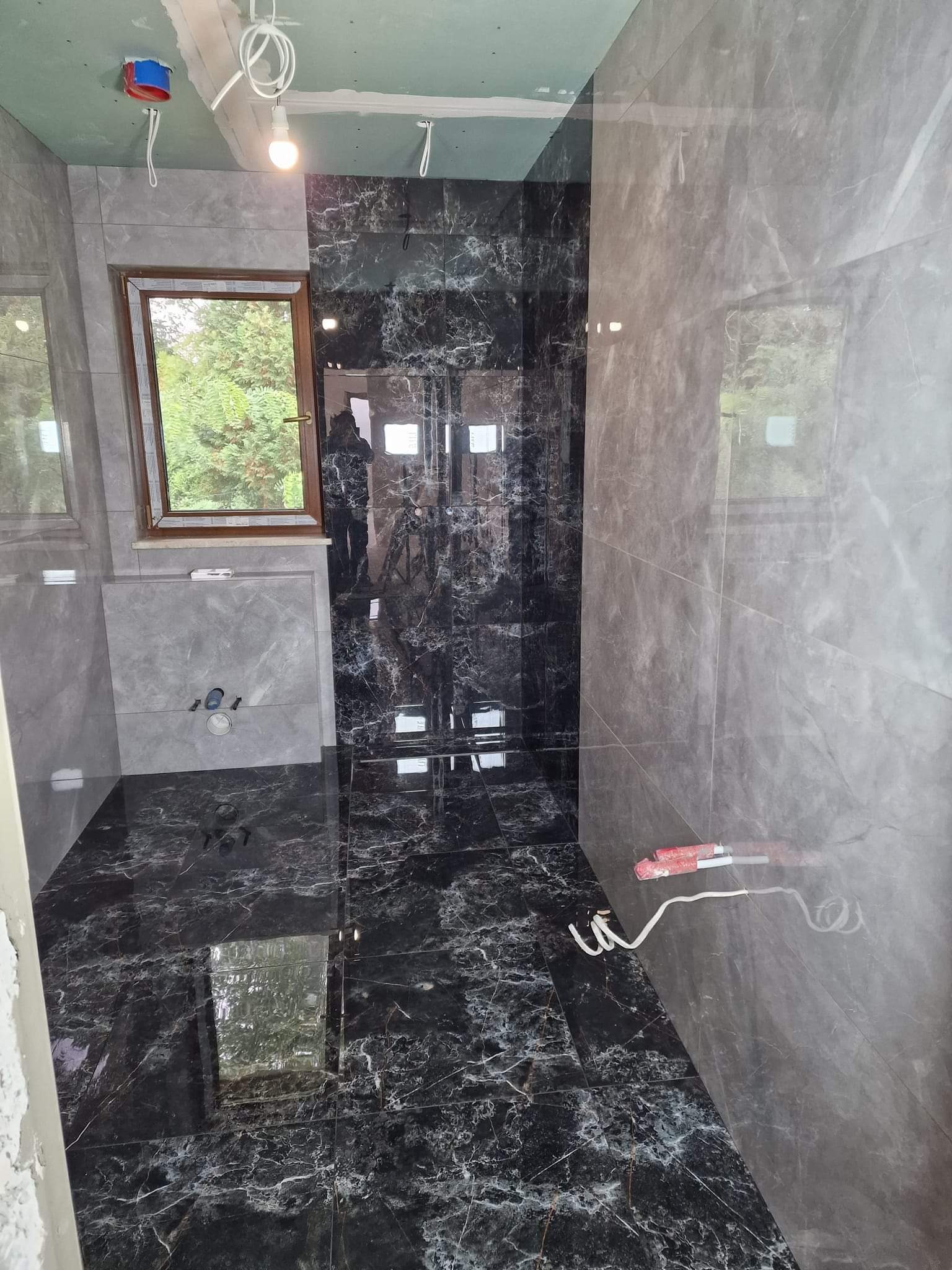 Nowoczesna łazienka w trakcie wykańczania: czarne marmurowe płytki na podłodze i ścianie prysznica, szare płytki na pozostałych ścianach, okno z widokiem na zieleń, widoczne instalacje elektryczne...