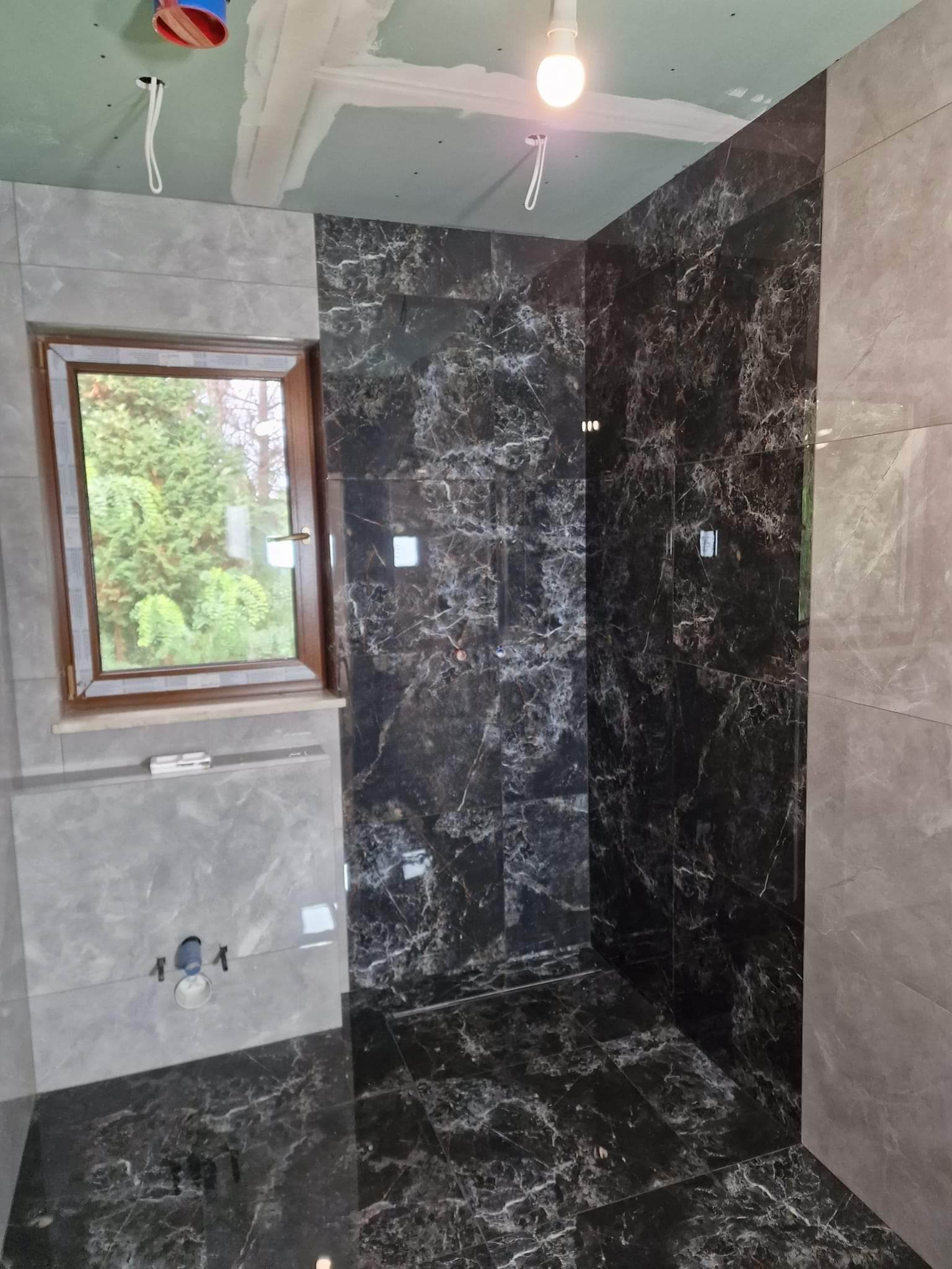 Wnętrze łazienki w trakcie prac wykończeniowych, widoczne płytki imitujące marmur w odcieniach szarości i czerni na podłodze i ścianach, okno z drewnianą ramą oraz surowy sufit z widocznymi...