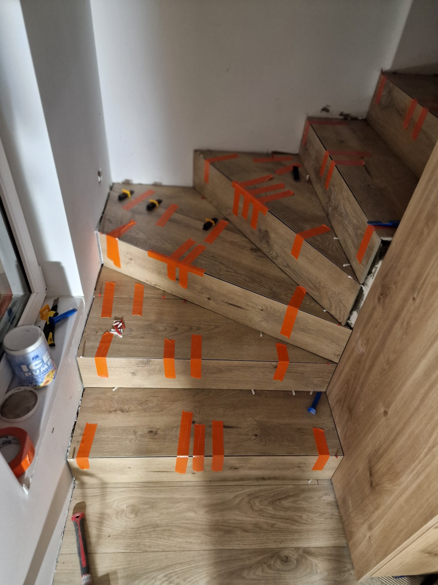 Montaż płytek imitujących drewno na schodach, zabezpieczonych pomarańczową taśmą, widoczne narzędzia i poziomice, etap prac wykończeniowych.