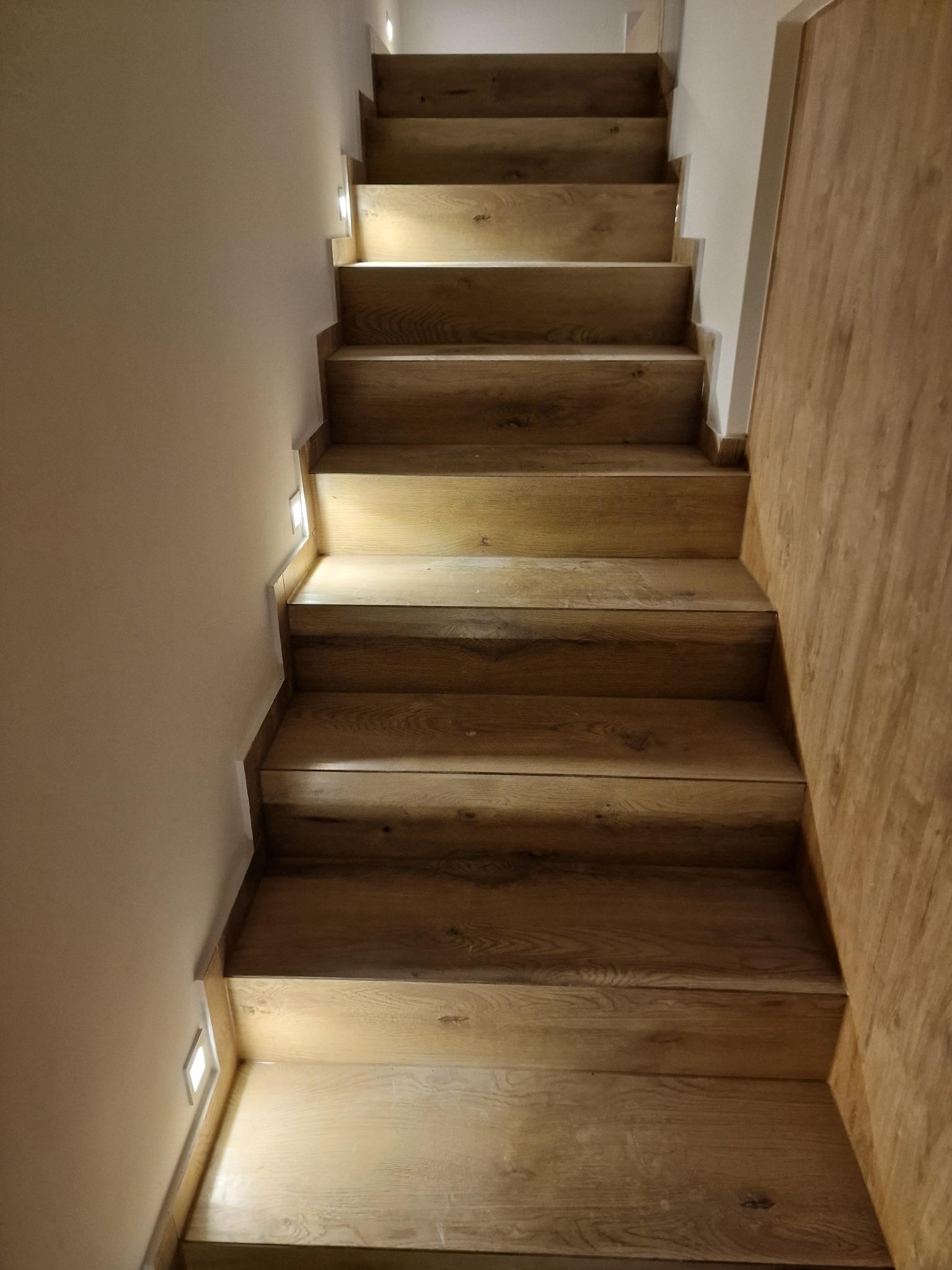 Drewniane schody z podświetleniem LED w ścianie, widok z dołu, jasne drewno z widocznymi słojami.