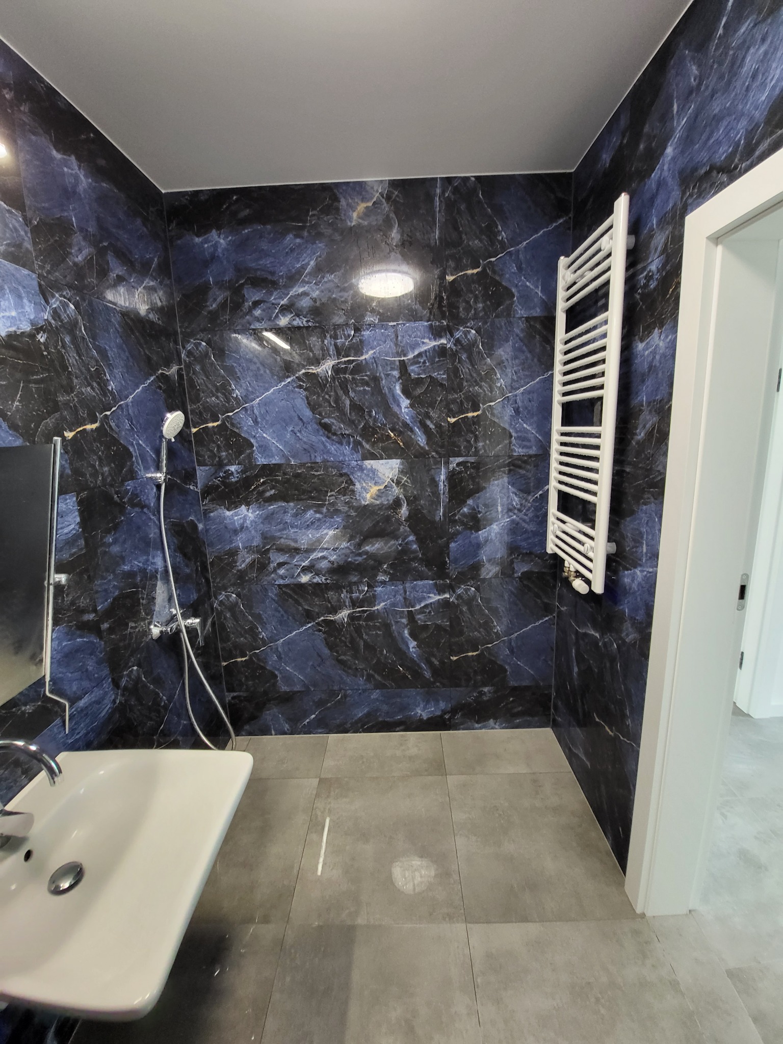 Wykończona łazienka z płytkami imitującymi czarny marmur z niebieskimi żyłami, biała umywalka, grzejnik drabinkowy i szara podłoga z płytek.