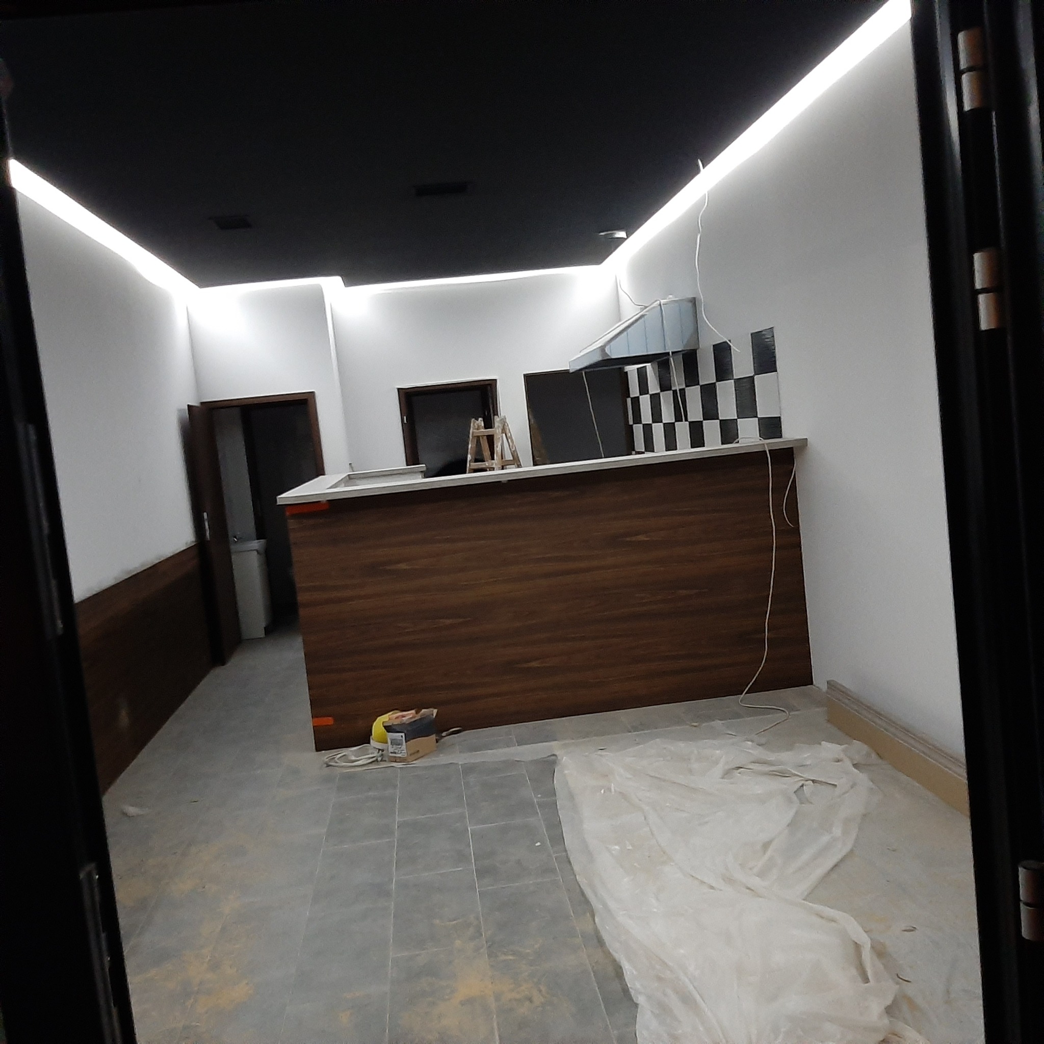 Wnętrze lokalu w trakcie prac wykończeniowych, widoczna drewniana zabudowa baru, czarno-białe płytki na ścianie, zabezpieczona folią podłoga i drabina w tle.