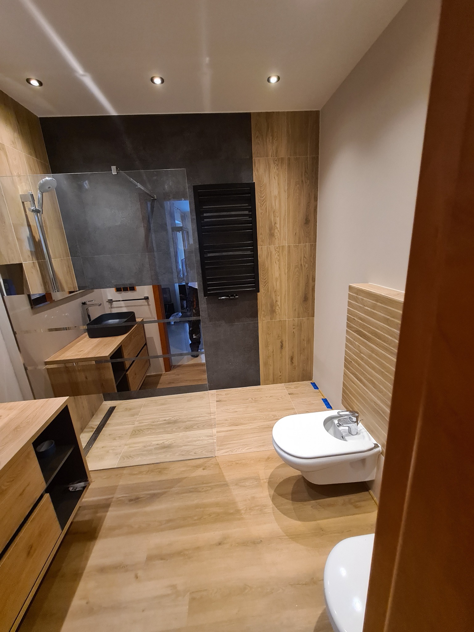 Nowoczesna łazienka z drewnopodobnymi płytkami, kabiną prysznicową typu walk-in i wiszącą miską WC oraz bidetem, oświetlona punktowymi lampami sufitowymi.