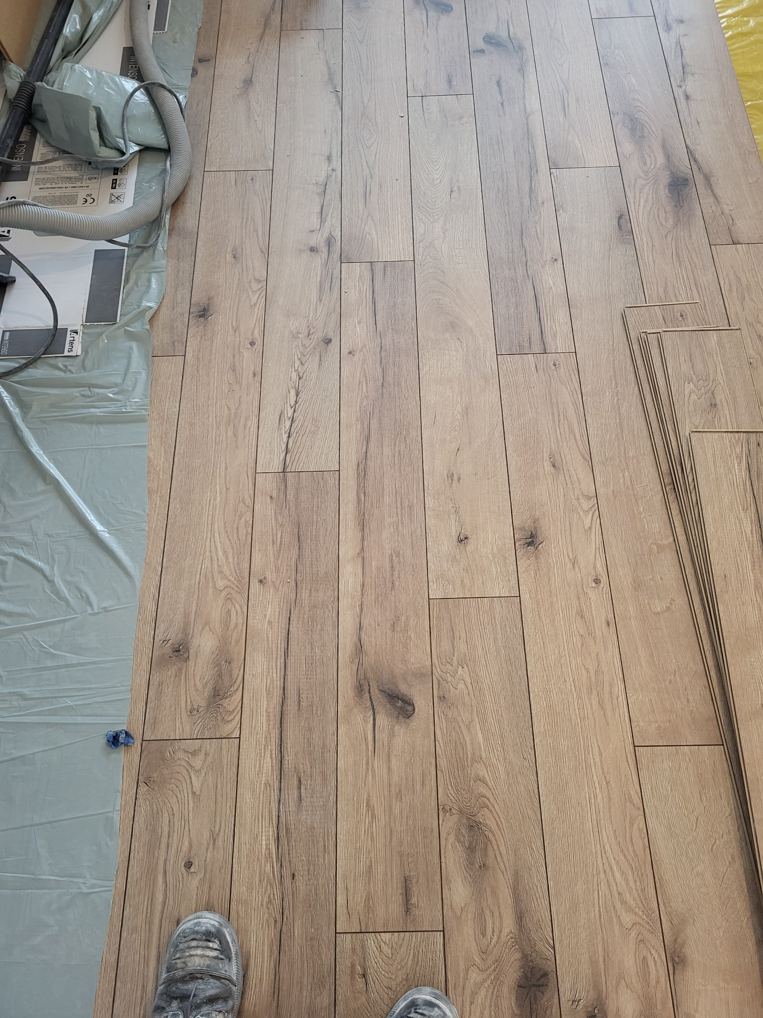 Układanie paneli podłogowych o wzorze drewna; widoczne panele czekające na montaż, zabezpieczona folią podłoga i fragment buta pracownika.