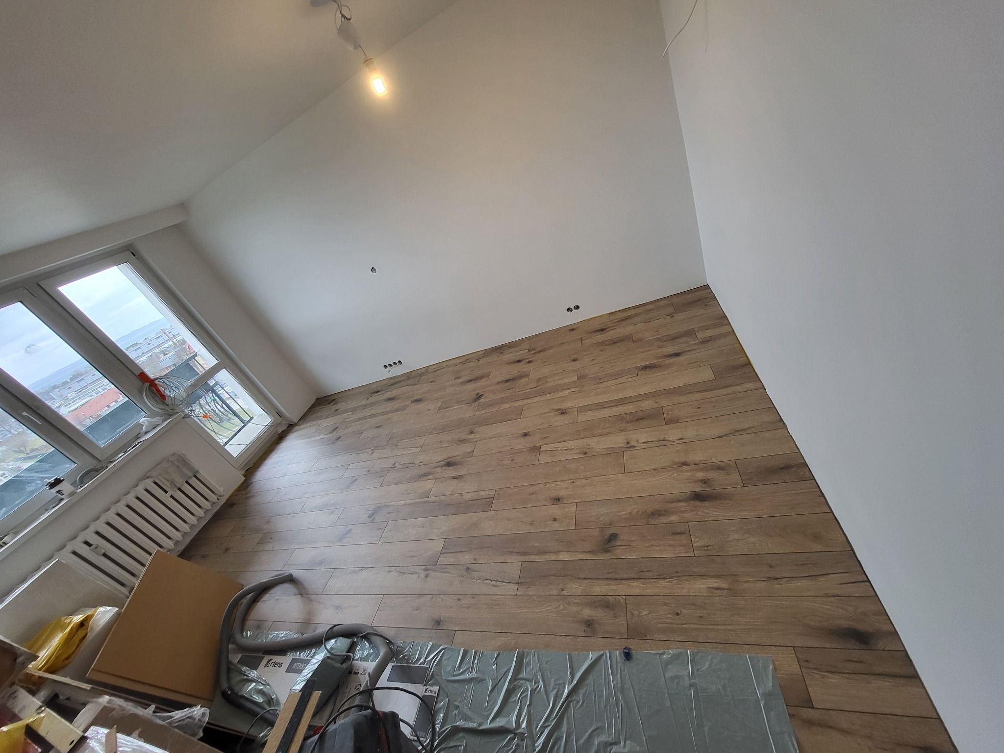 Pokój po remoncie: Świeżo położona podłoga z paneli drewnopodobnych, białe ściany i sufit, okno z widokiem na miasto. Na podłodze folia ochronna i sprzęt budowlany.