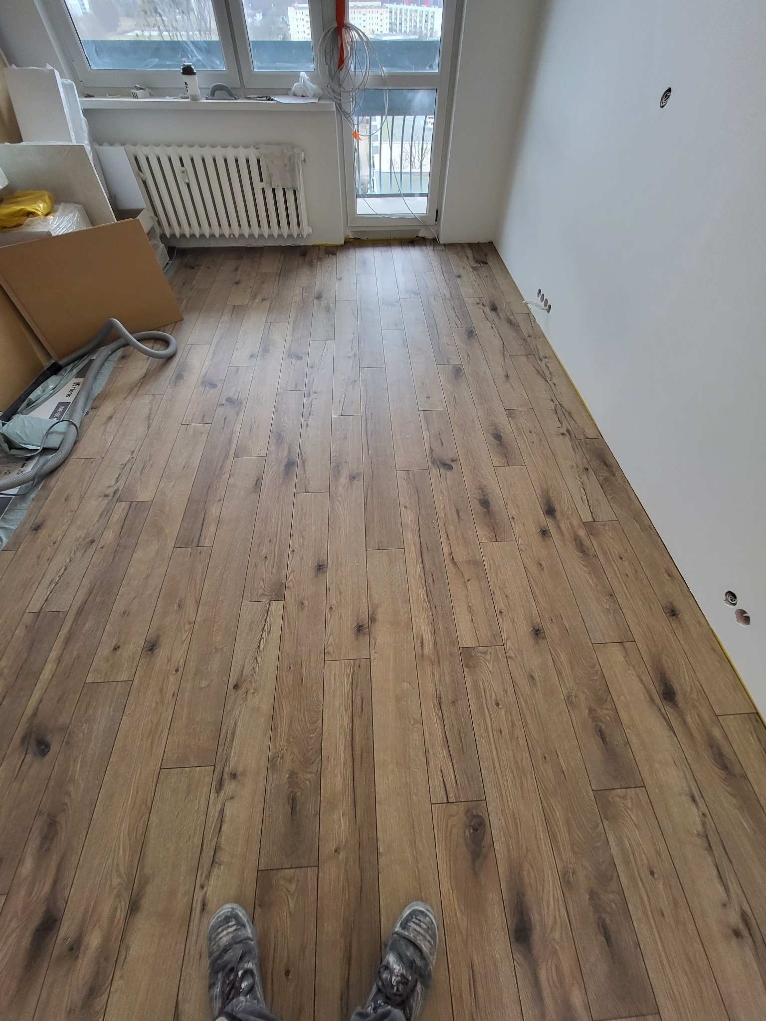 Ułożona podłoga z paneli drewnopodobnych w jasnym odcieniu brązu, widoczne gniazdka elektryczne w ścianie i fragmenty materiałów budowlanych przy oknie.