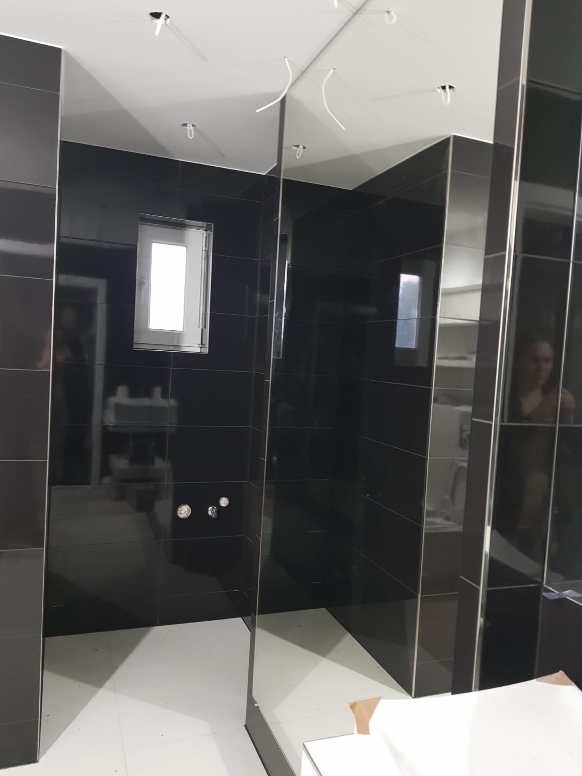 Czarne, błyszczące płytki ścienne w łazience, biała podłoga, widoczne przyłącza wodne, surowe wykończenie sufitu z wystającymi kablami, małe okno z białymi ramami.