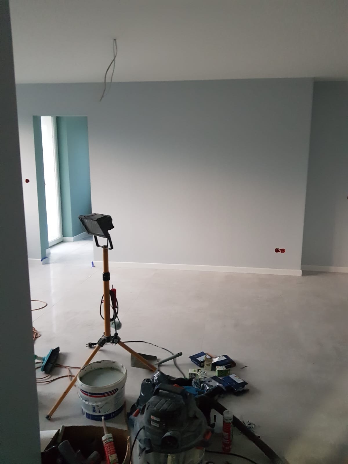 Pokój w trakcie odświeżania, świeżo pomalowane ściany na jasny odcień szarości, widoczny reflektor na statywie, wiadro z farbą i narzędzia malarskie na podłodze, wystające kable ze ściany.