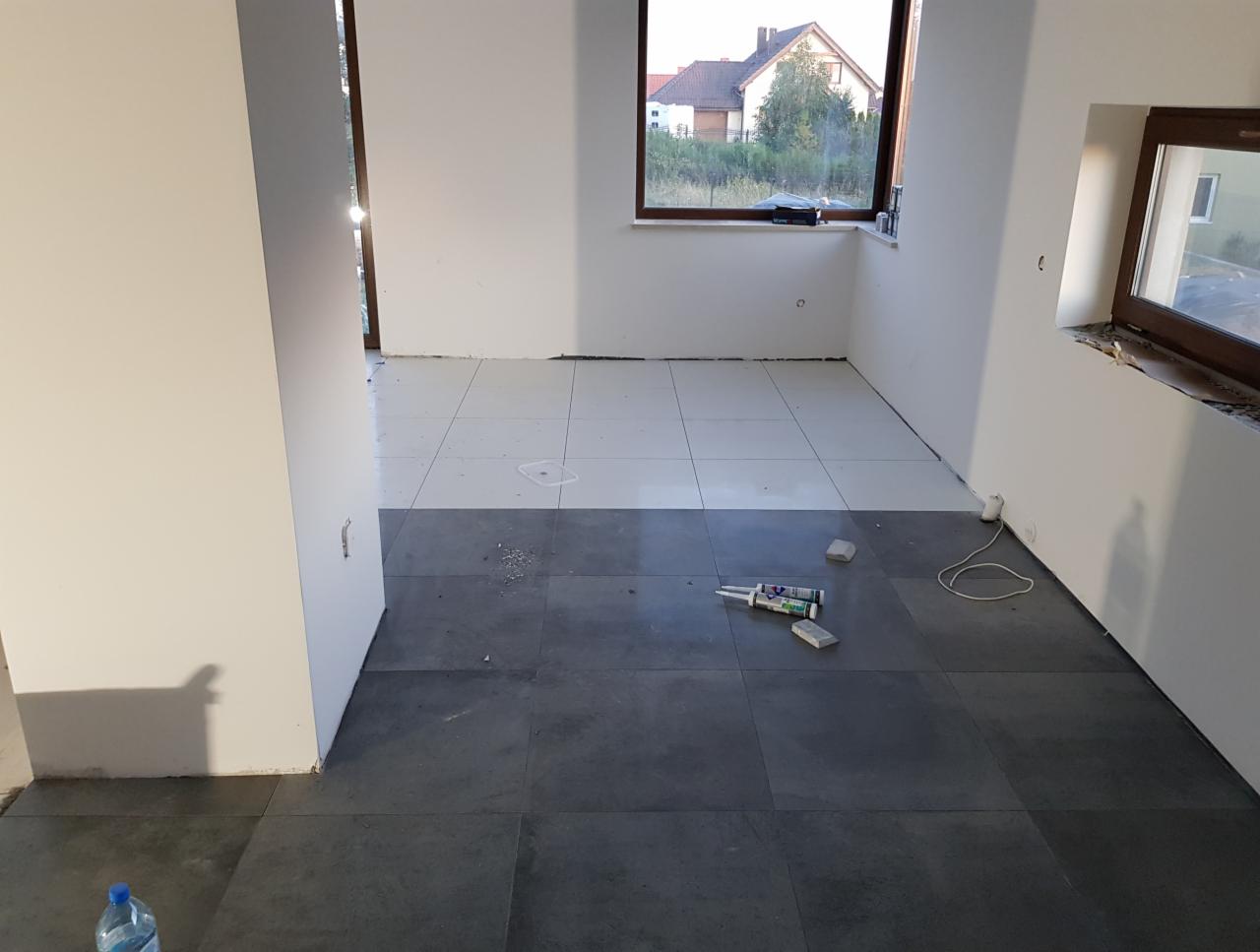 Wnętrze pokoju z częściowo ułożoną podłogą z szarych i białych płytek, widoczne narzędzia glazurnicze, okna z drewnianymi ramami, białe ściany w trakcie wykończenia.